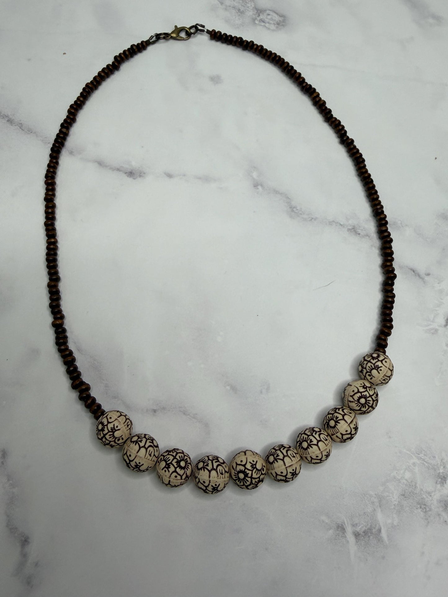 A beaded necklace featuring a strand of dark brown beads and a row of larger cream-colored beads with black crackle patterns, displayed on a white marble surface.