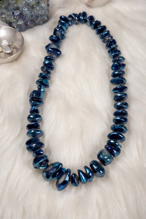 Blue beaded quartz necklace on a white surface with a stone and small dish in the background
