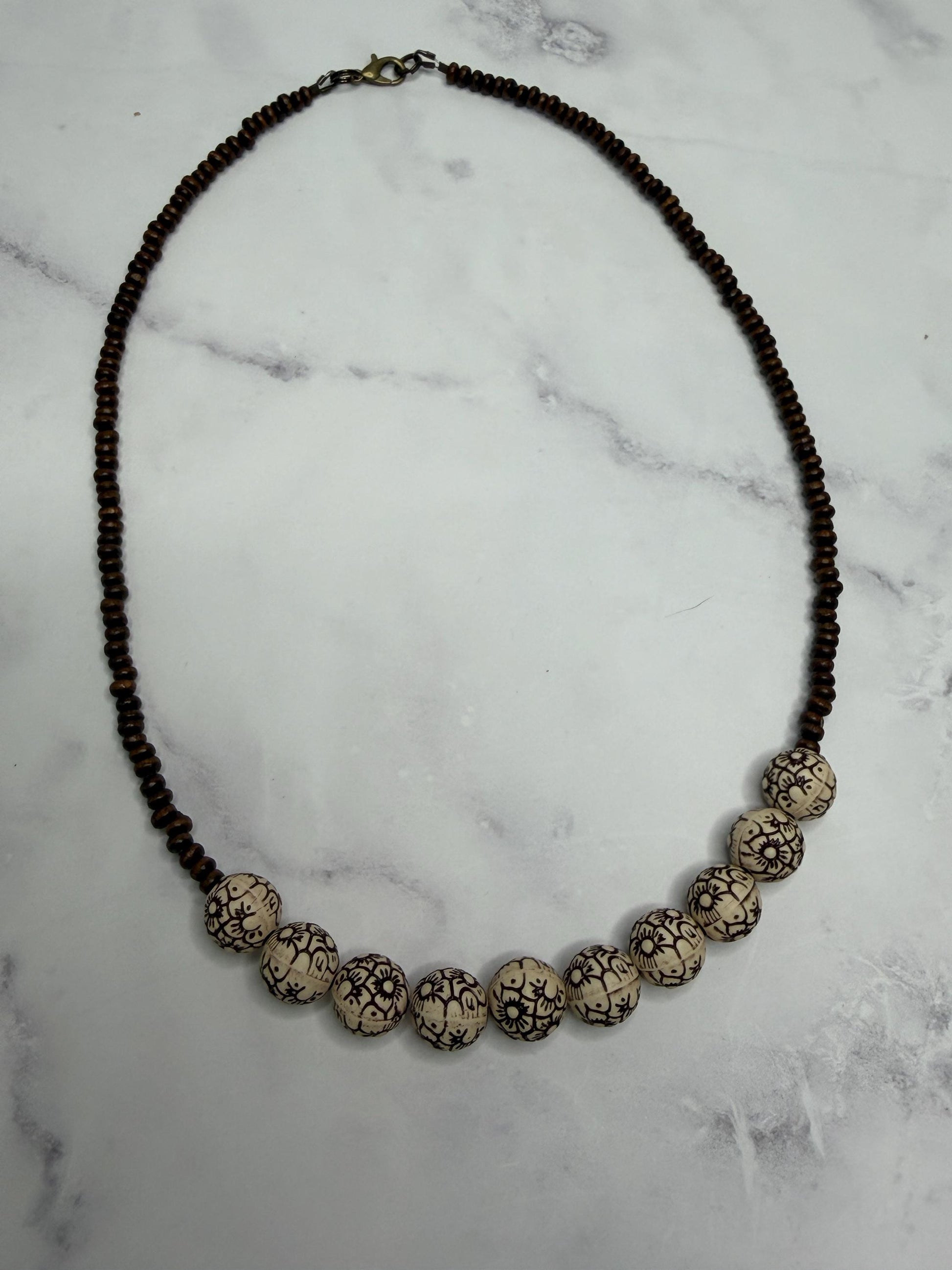 A beaded necklace featuring a strand of dark brown beads and a row of larger cream-colored beads with black crackle patterns, displayed on a white marble surface.