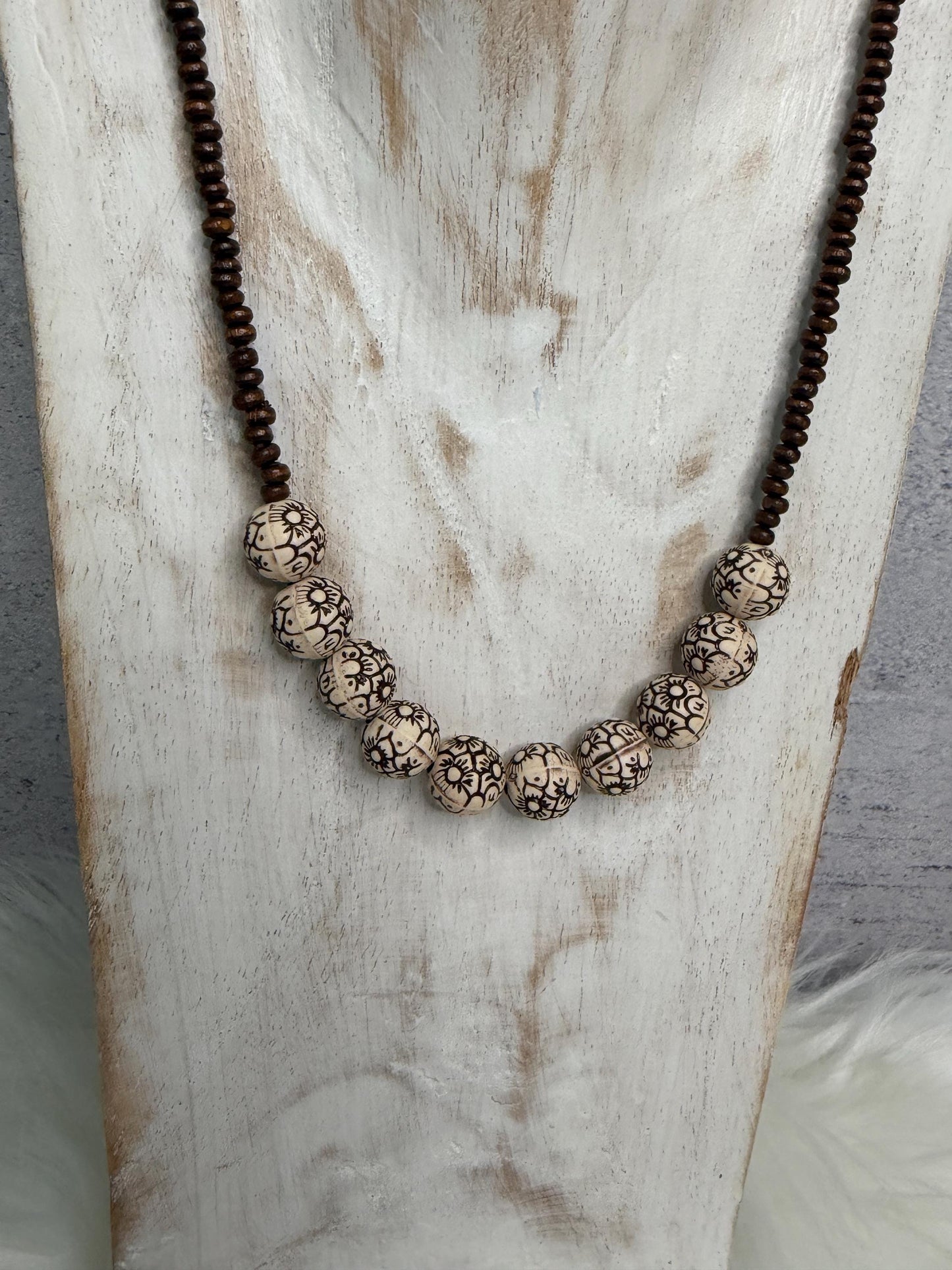 A beaded necklace displayed on a rustic white wooden stand. The necklace features a dark brown beaded chain with a central cluster of round, textured cream beads with black organic patterns.