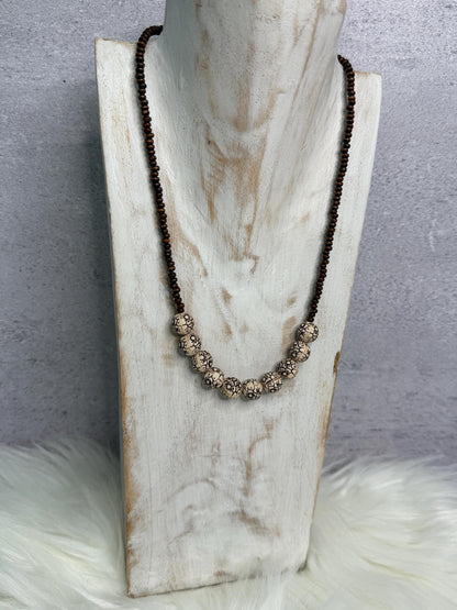 A long beaded necklace displayed on a distressed white wooden bust. The necklace features dark brown beads on the upper section and a row of larger textured metallic silver beads on the lower center.