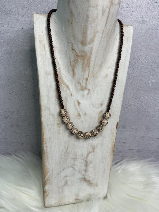 A long beaded necklace displayed on a distressed white wooden bust. The necklace features dark brown beads on the upper section and a row of larger textured metallic silver beads on the lower center.