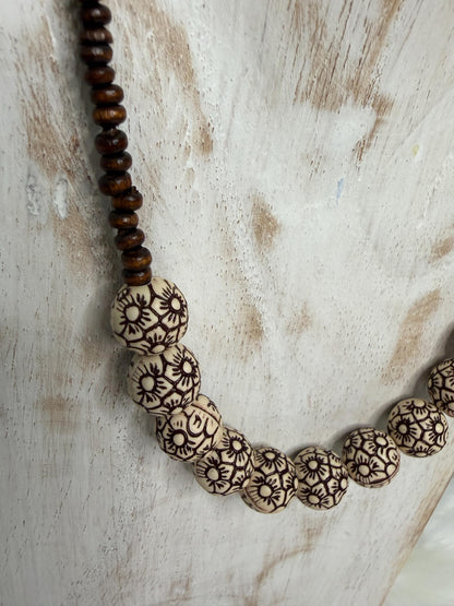 A close-up of a beaded necklace draped over a distressed white wooden surface. The necklace features a strand of dark brown wooden beads at the top transitioning into large, round cream-colored beads with intricate dark brown floral or sunburst engravings.