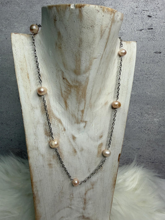 A necklace with irregular light pink pearls spaced along a dark oxidized chain, displayed on a distressed wood neck stand against a gray textured background with white faux fur accents.