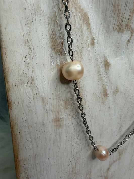 A close-up of a necklace with two large, irregular cream-colored freshwater pearls on a dark oxidized metal chain, draped against a distressed white wood background.